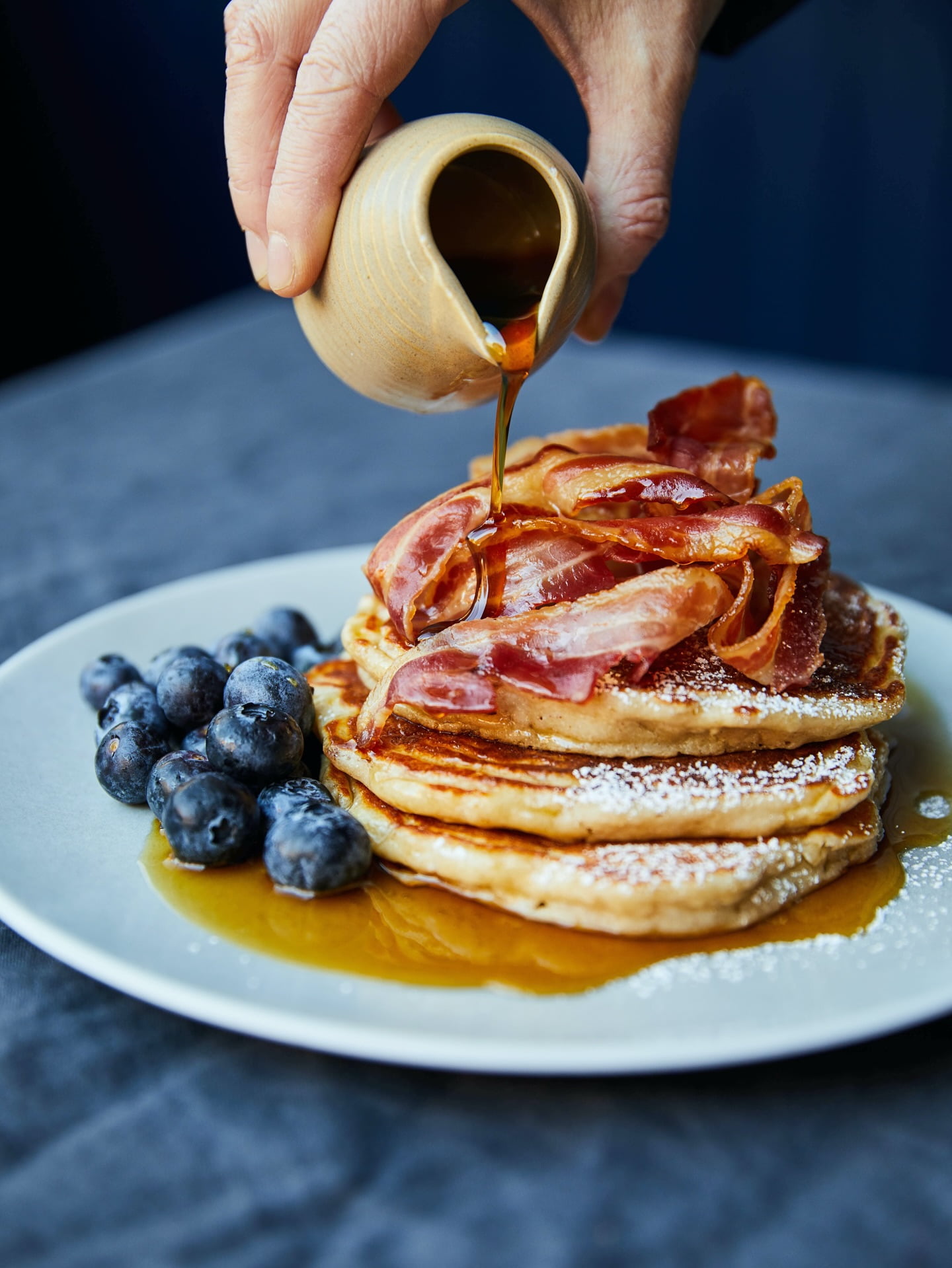 A hand pours maple syrup over a stack of three pancakes topped with crispy bacon next to fresh blueberries on a grey plate.